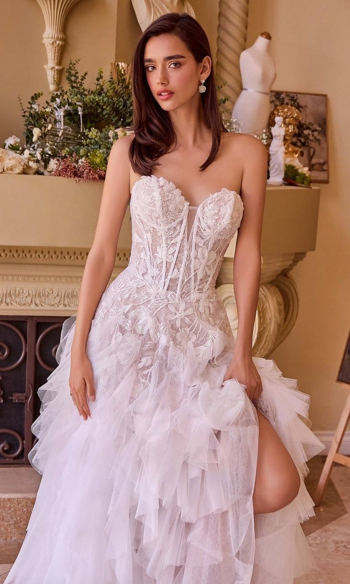 A woman in the Andrea and Leo WL016 long wedding dress, featuring a strapless lace-applique corset bodice and ruffled tulle A-line skirt, stands indoors near a decorated mantelpiece.